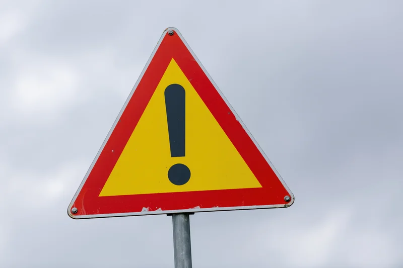 Triangular warning road sign with exclamation mark on metal pole against cloudy sky