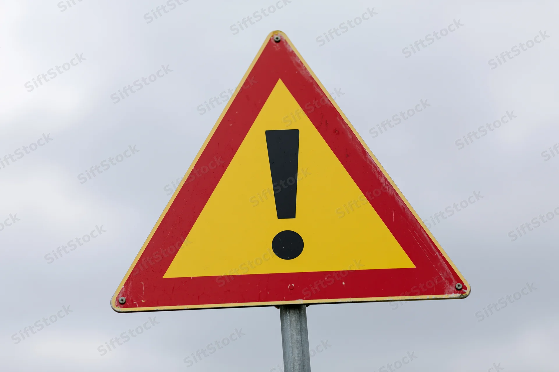 Triangular warning road sign with black exclamation mark on red and yellow background mounted on metal pole against cloudy sky