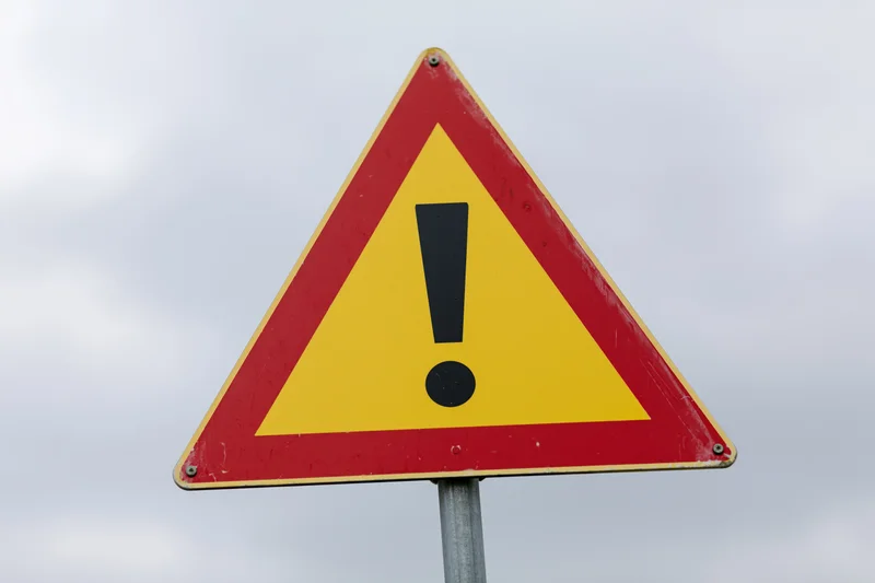 Triangular warning road sign with black exclamation mark on red and yellow background mounted on metal pole against cloudy sky