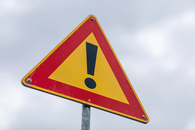 Triangular warning road sign with black exclamation mark on red and yellow background mounted on metal post against cloudy sky