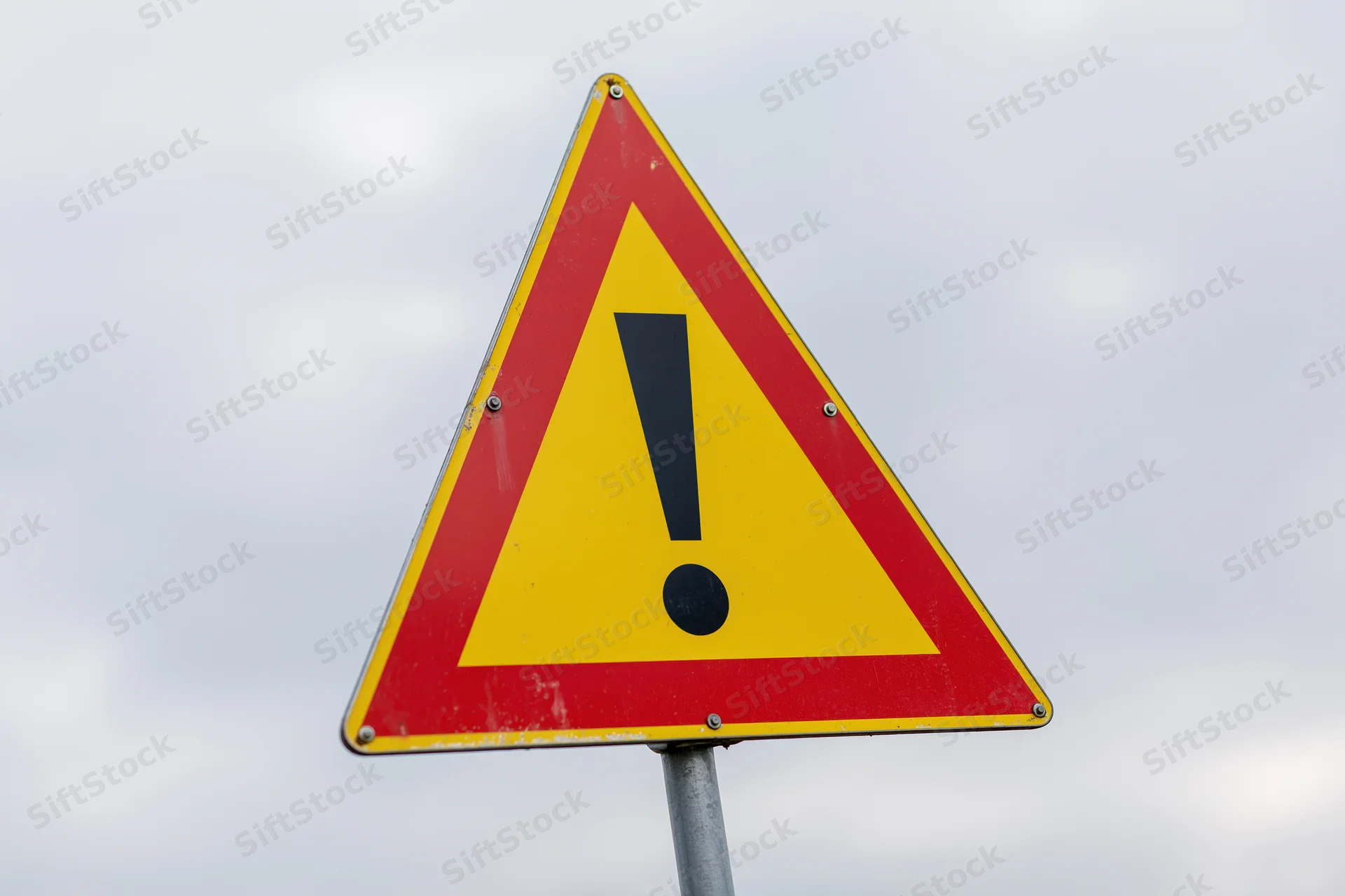 Triangular warning road sign with black exclamation mark on yellow and red background against cloudy sky