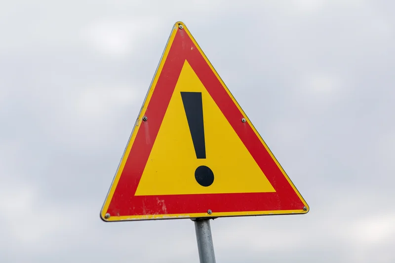 Triangular warning road sign with black exclamation mark on yellow and red background against cloudy sky