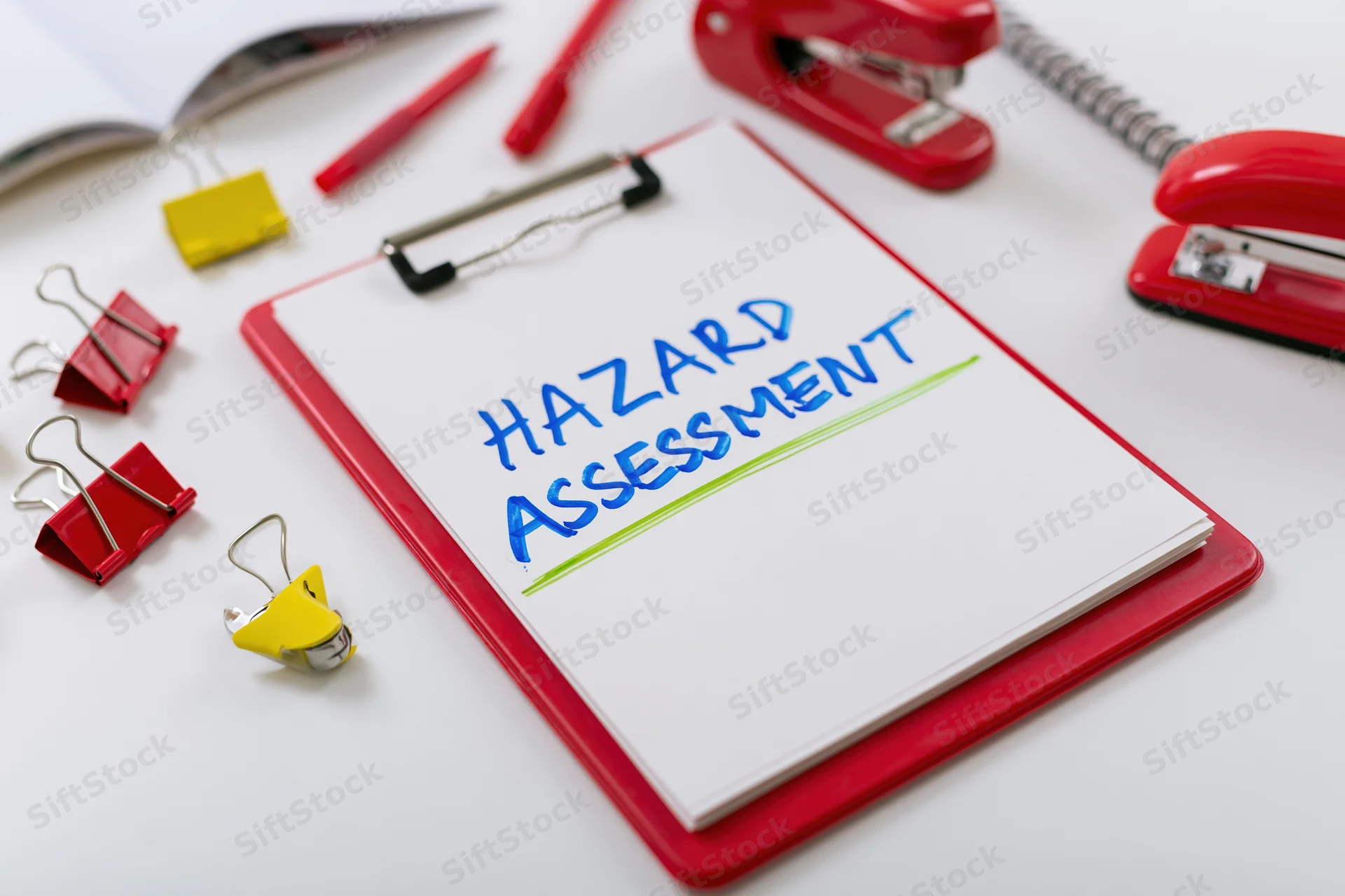 Red clipboard with white paper labeled hazard assessment on a desk surrounded by office supplies