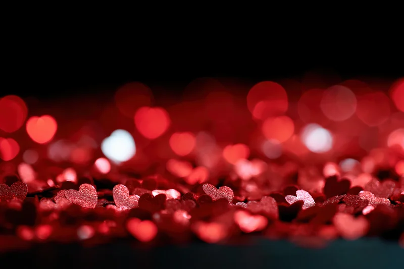 Red heart-shaped glitter scattered across a dark surface with bokeh lights in the background