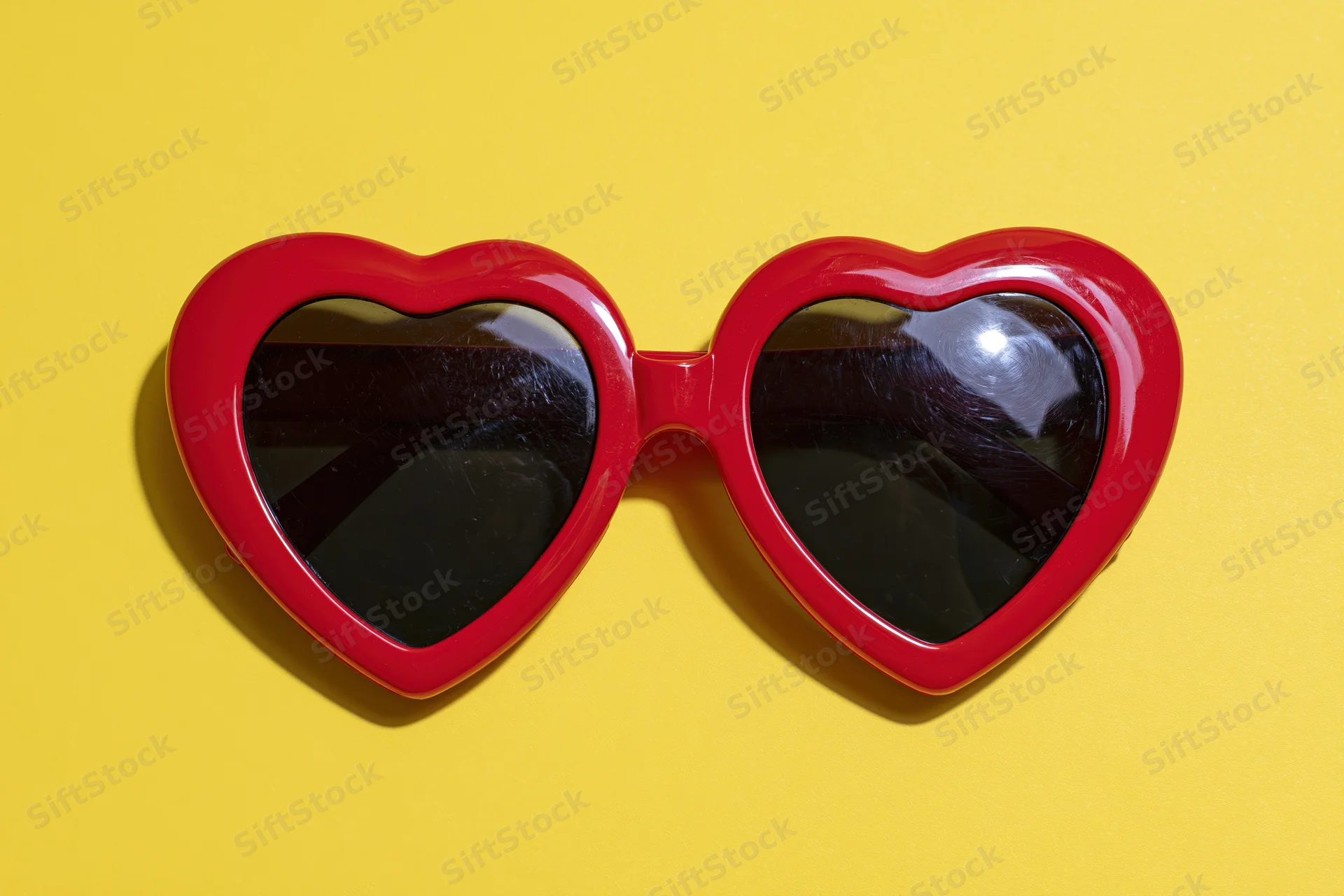 Red heart-shaped sunglasses with glossy plastic frames lying on yellow background