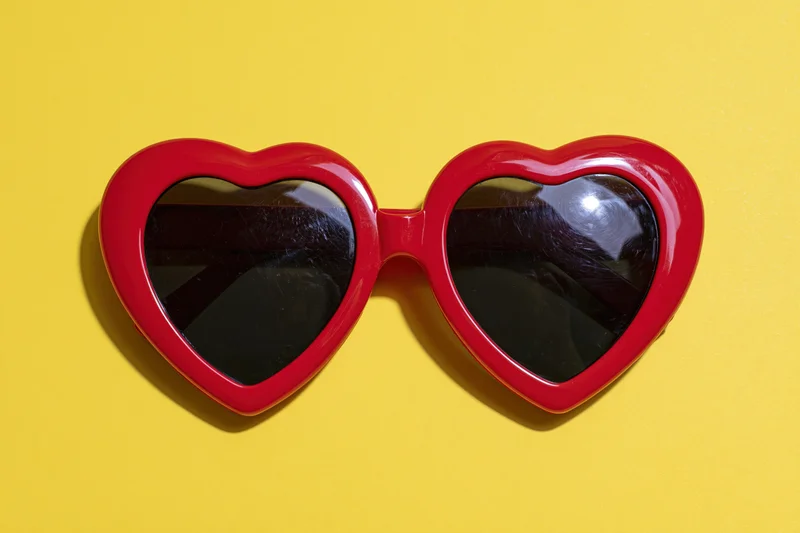 Red heart-shaped sunglasses with glossy plastic frames lying on yellow background