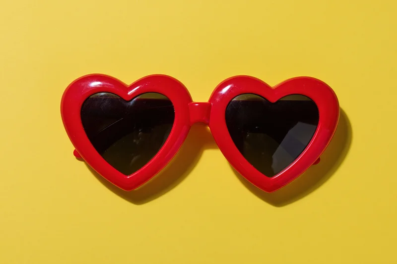 Red heart-shaped sunglasses with glossy plastic frames lying on a yellow background