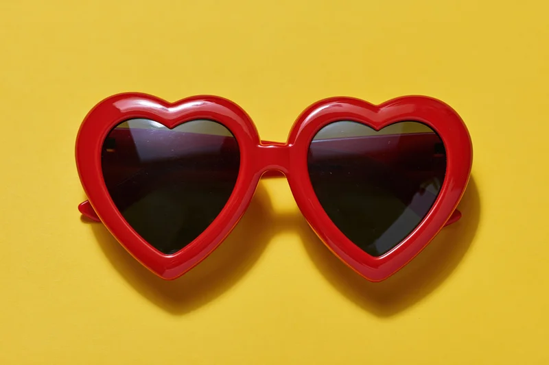 Red heart-shaped sunglasses with glossy plastic frames lying on yellow background