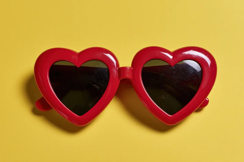 Red heart-shaped sunglasses with glossy plastic frames lying on yellow background