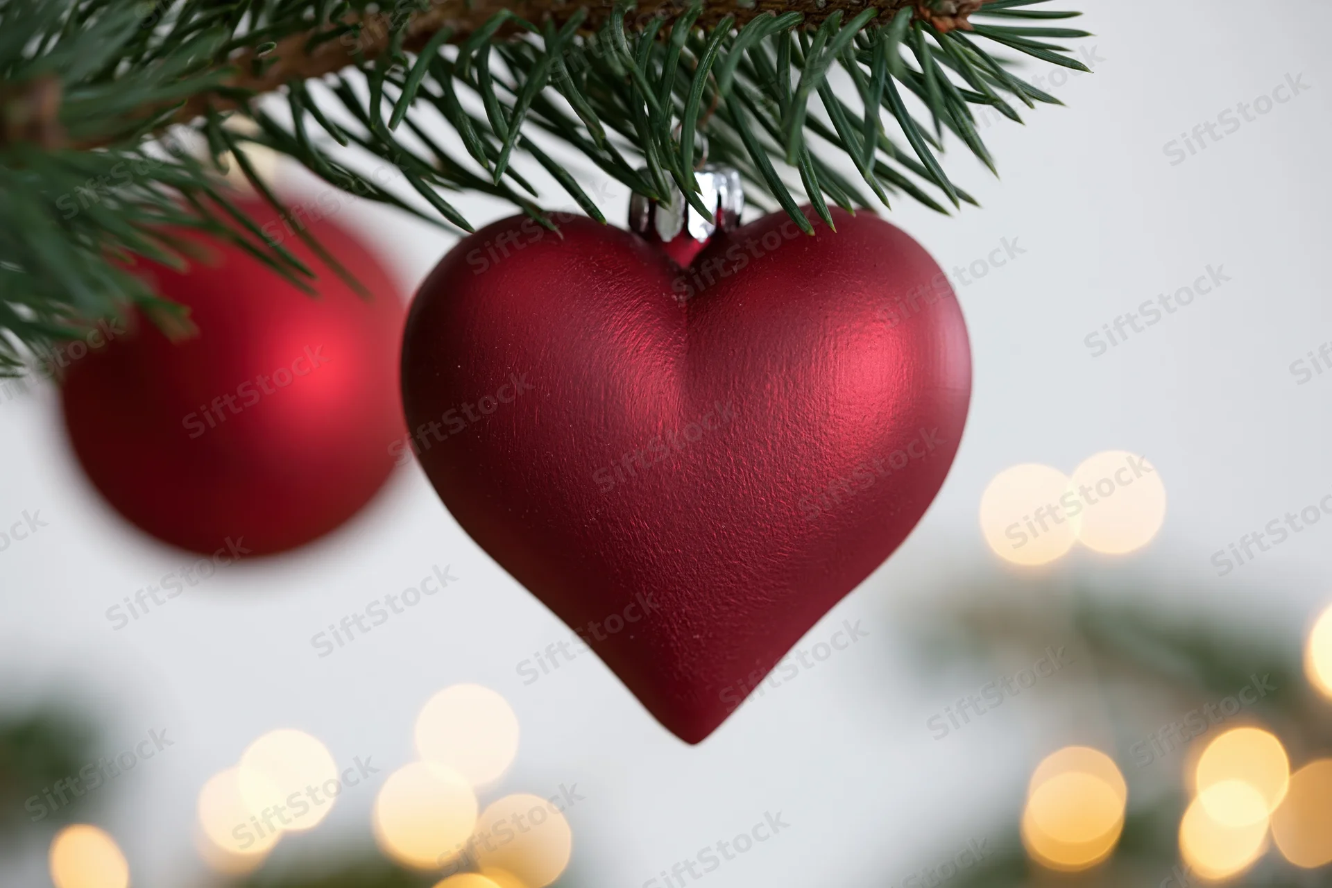 Red matte heart-shaped ornament hanging from pine tree branch with blurred lights in background
