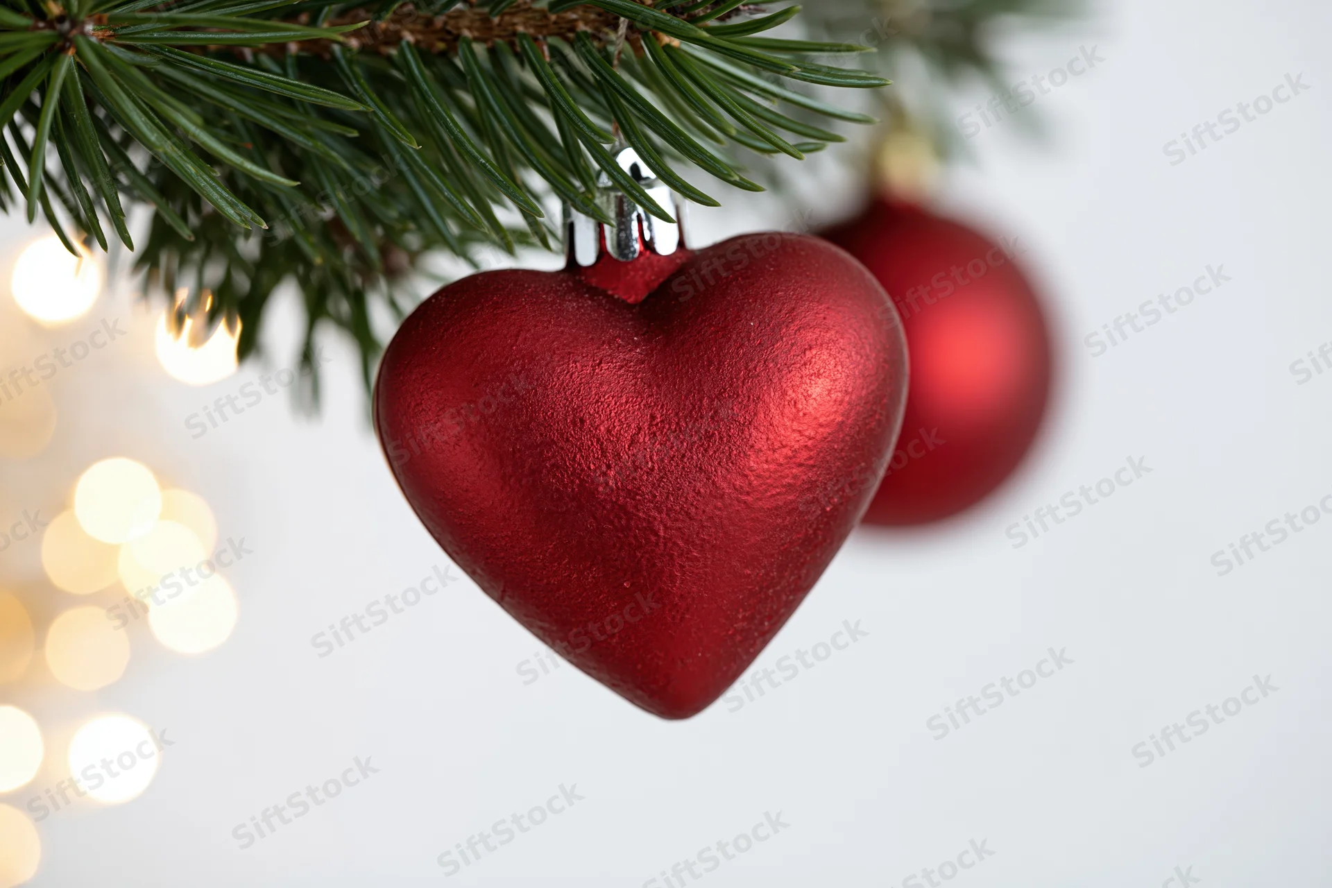 Red heart-shaped ornament hanging from a pine tree branch with blurred lights in the background