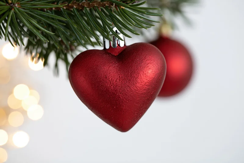 Red heart-shaped ornament hanging from a pine tree branch with blurred lights in the background
