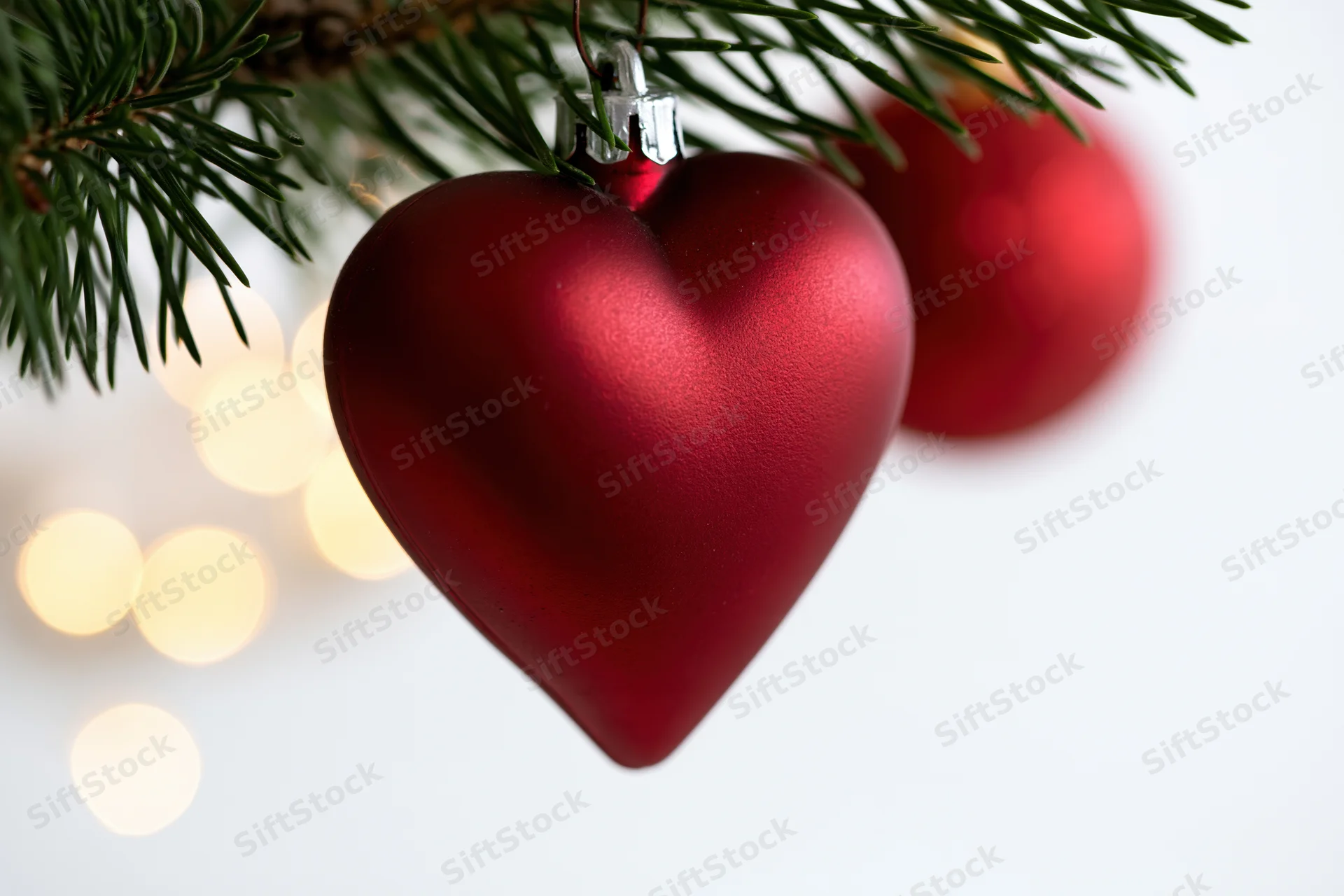 Red heart-shaped christmas ornament hanging from pine tree branch with blurred lights in background
