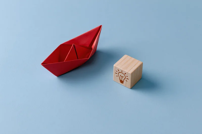 Red origami paper boat moving toward a wooden block with lightbulb symbol on blue surface