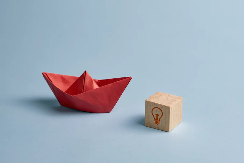 Red origami paper boat next to wooden block with lightbulb symbol on flat blue surface