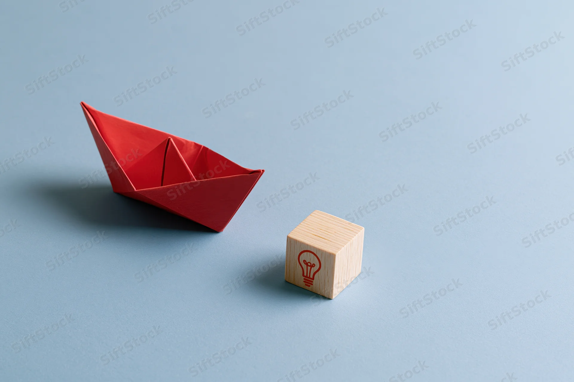 Red origami paper boat moving toward a wooden block with lightbulb symbol on blue surface