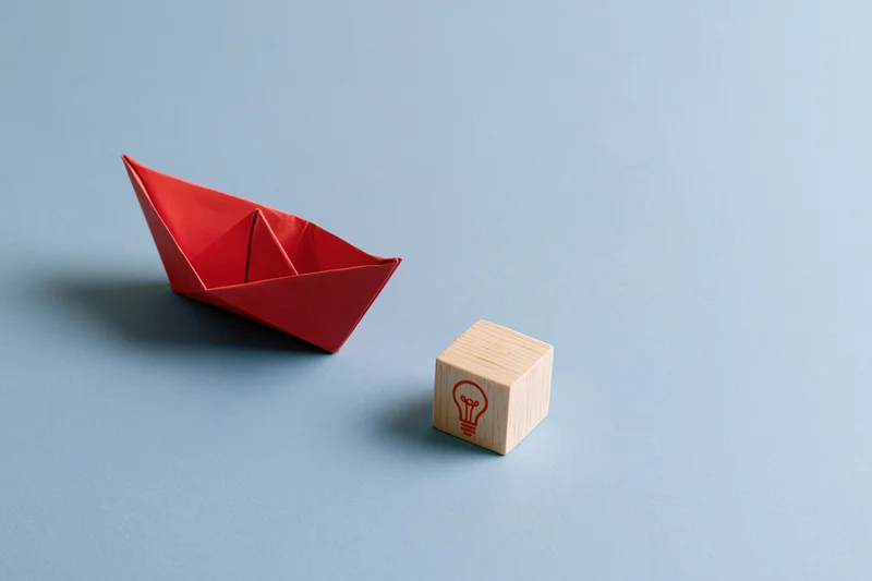 Red origami paper boat moving toward a wooden block with lightbulb symbol on blue surface