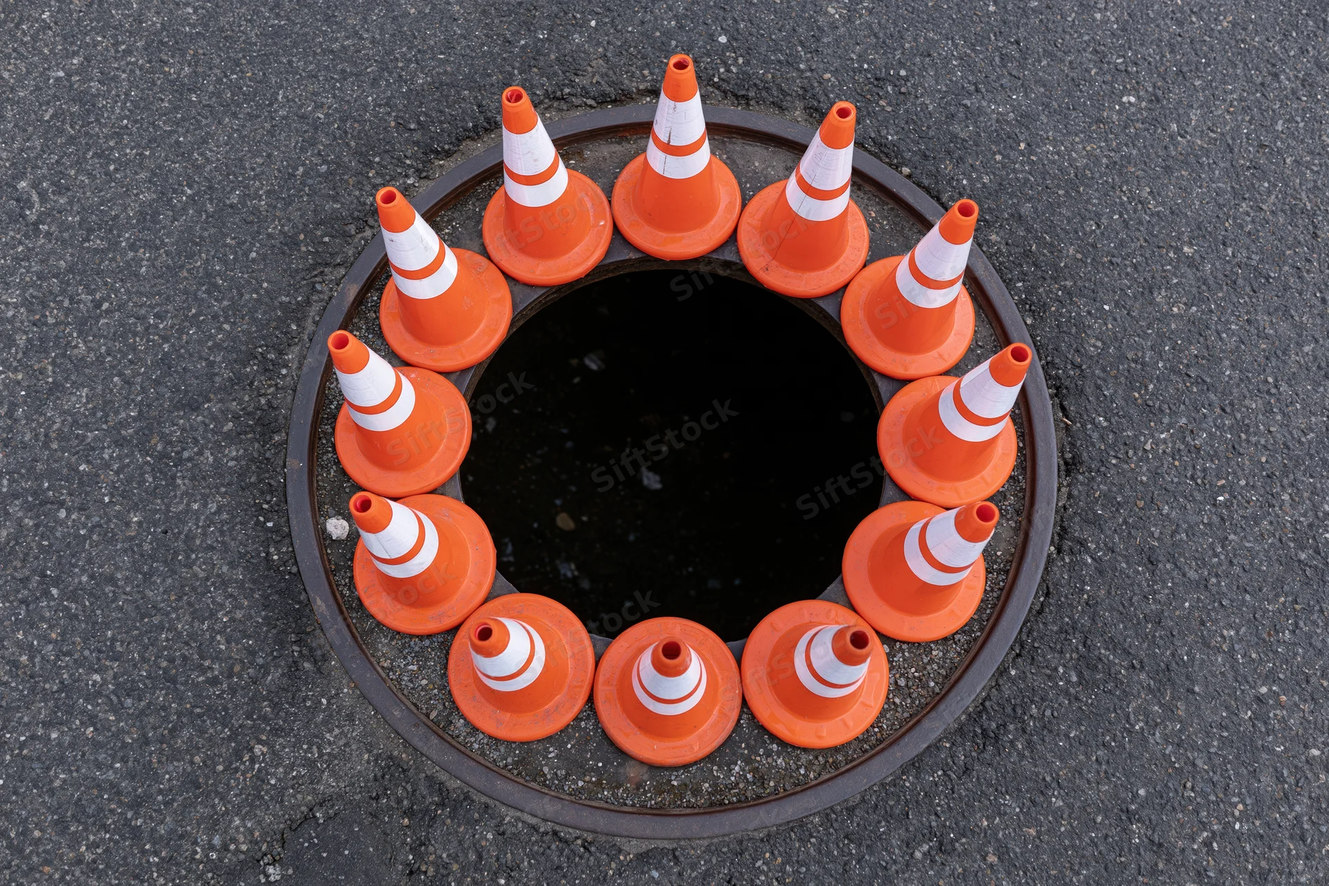 Small orange and white traffic cones arranged in a circle around an open manhole on asphalt road