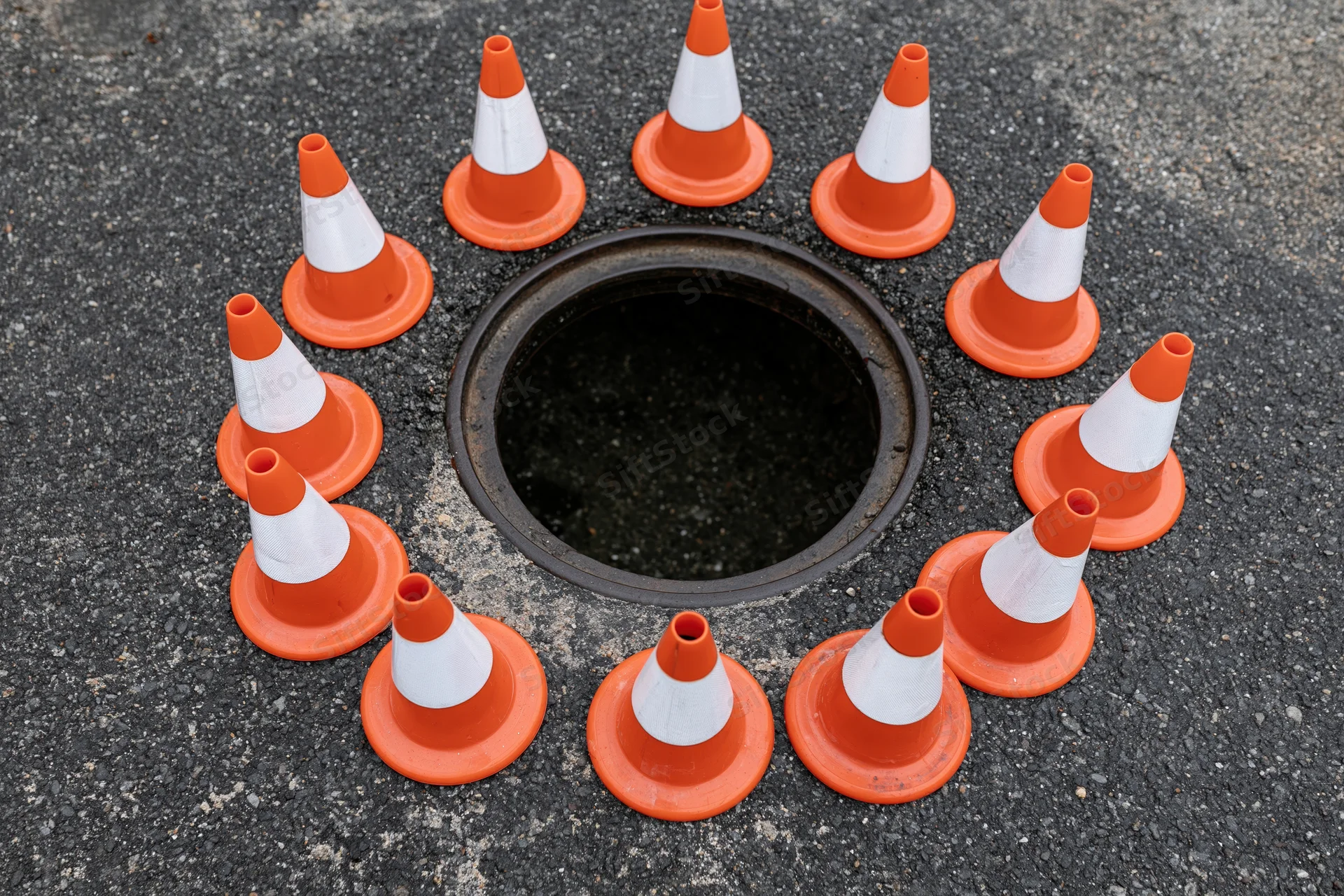 Small orange and white traffic cones arranged in a circle around an open manhole on asphalt pavement
