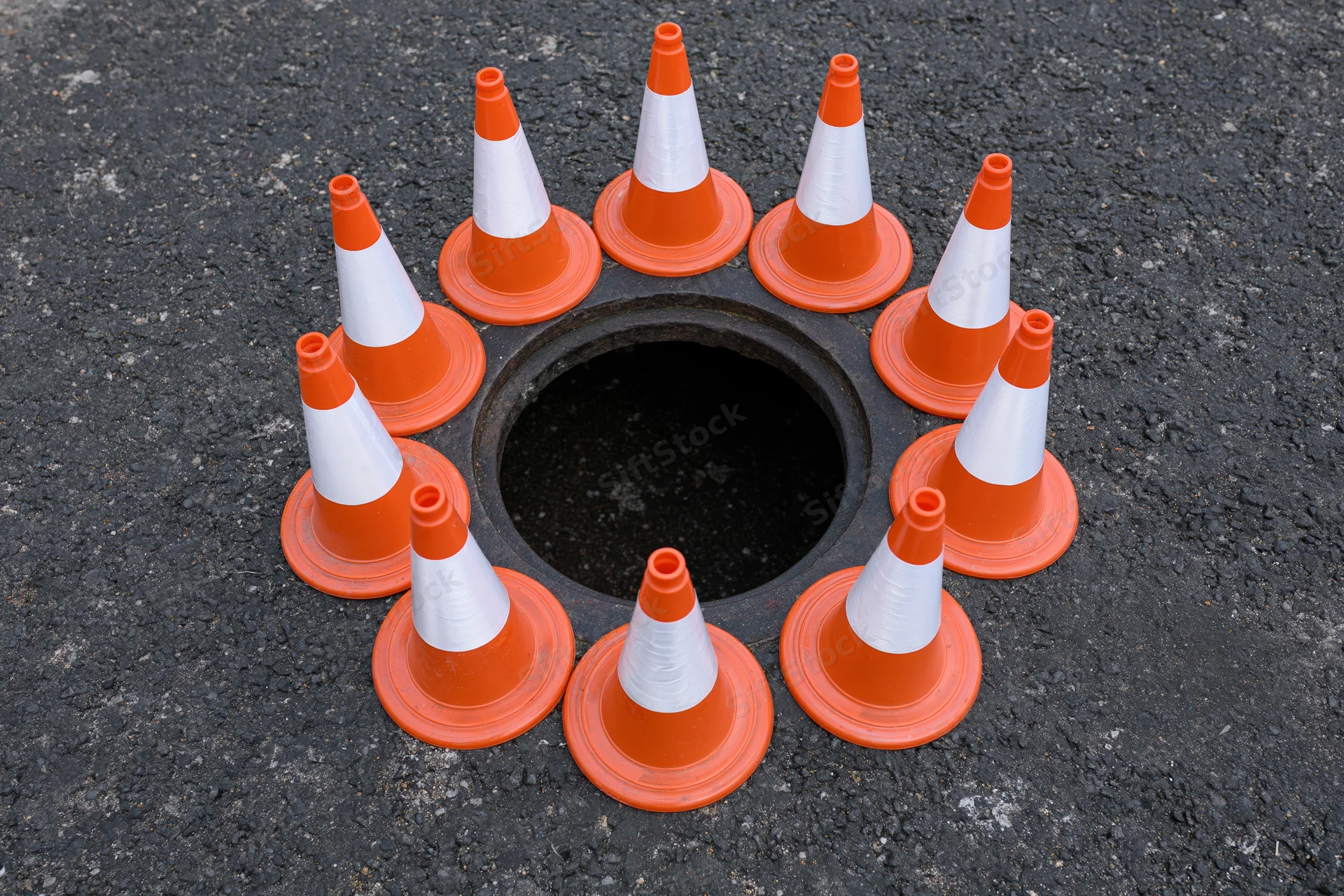 Small orange and white traffic cones arranged in a circle around an open manhole on asphalt road