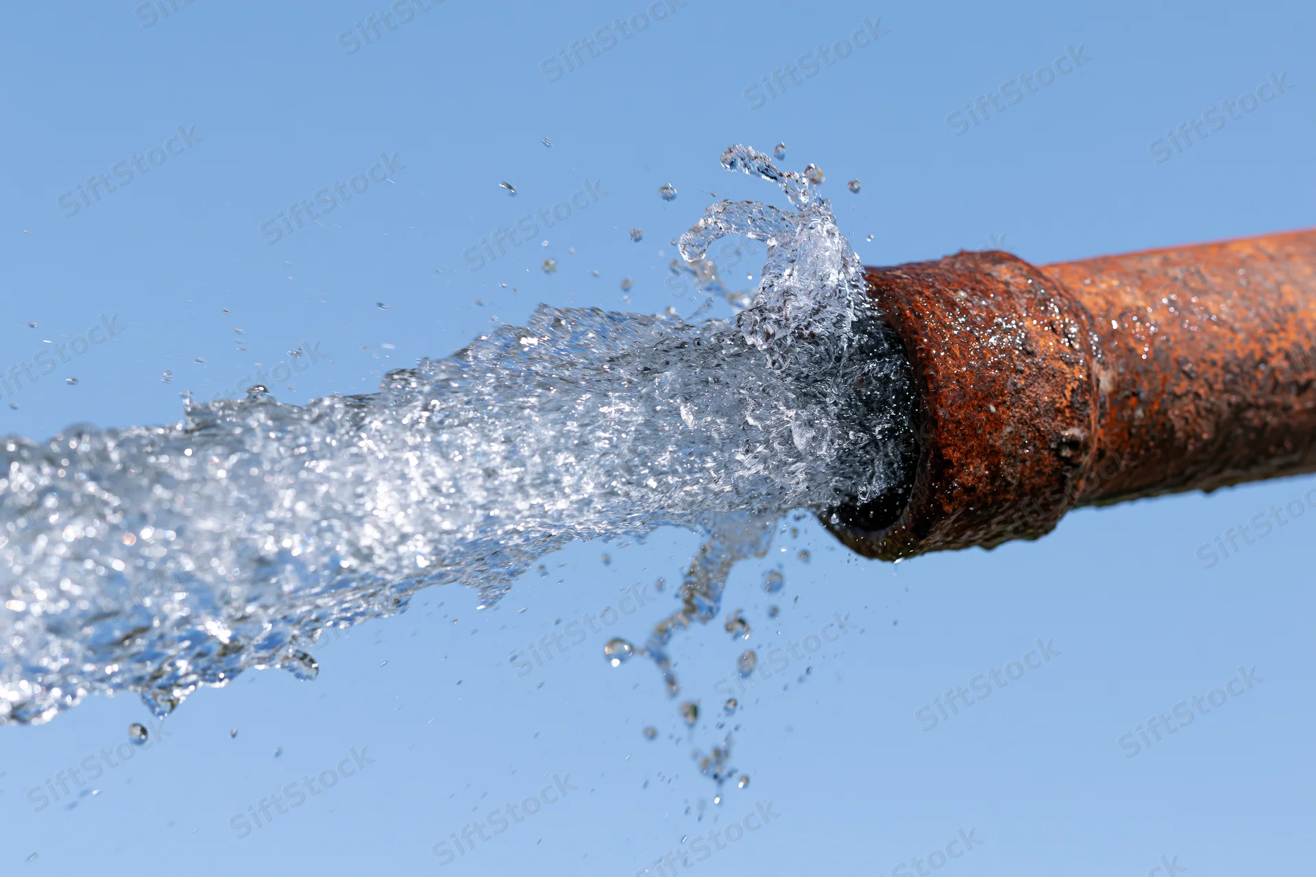 Rusty metal pipe bursting with high-pressure water spray against blue sky