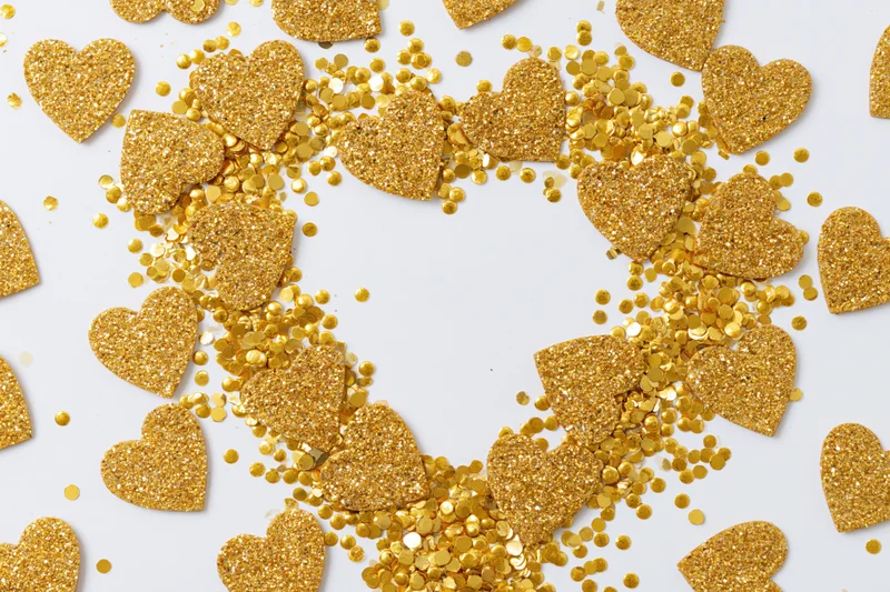 Gold glitter hearts and round confetti scattered on white surface forming a heart shape with copyspace in center