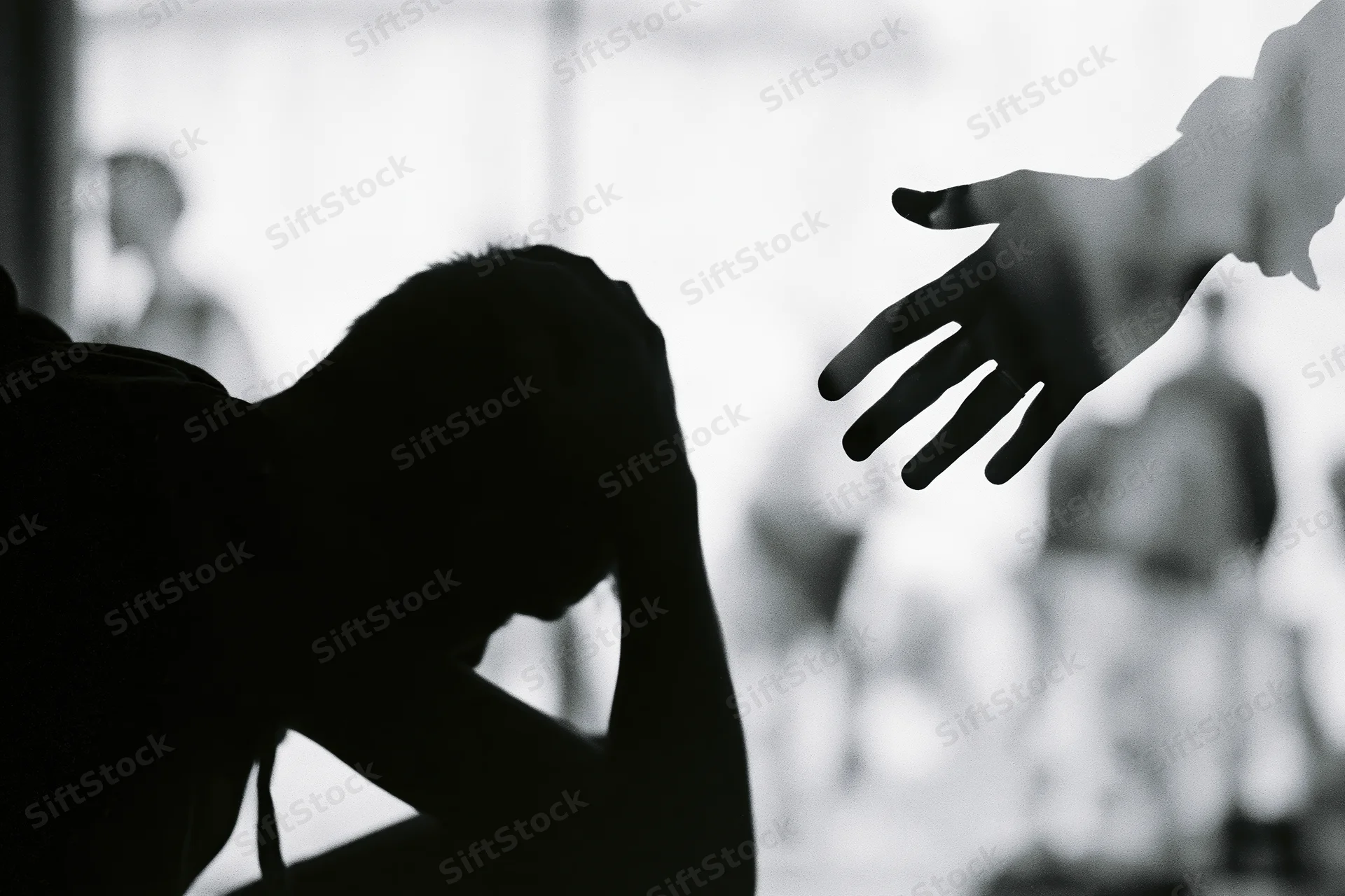 Silhouette of a distressed young person hunched with head in hands as a hand reaches toward them in a therapy room