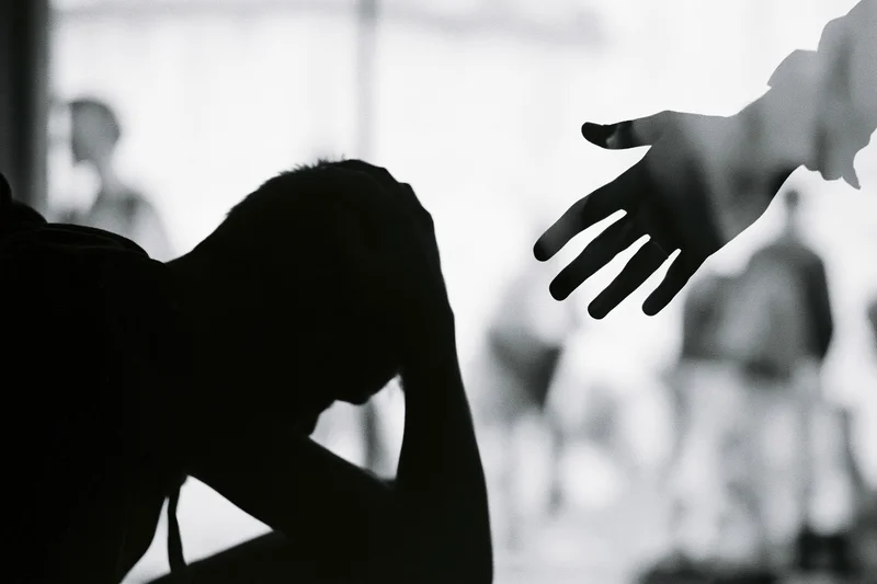 Silhouette of a distressed young person hunched with head in hands as a hand reaches toward them in a therapy room