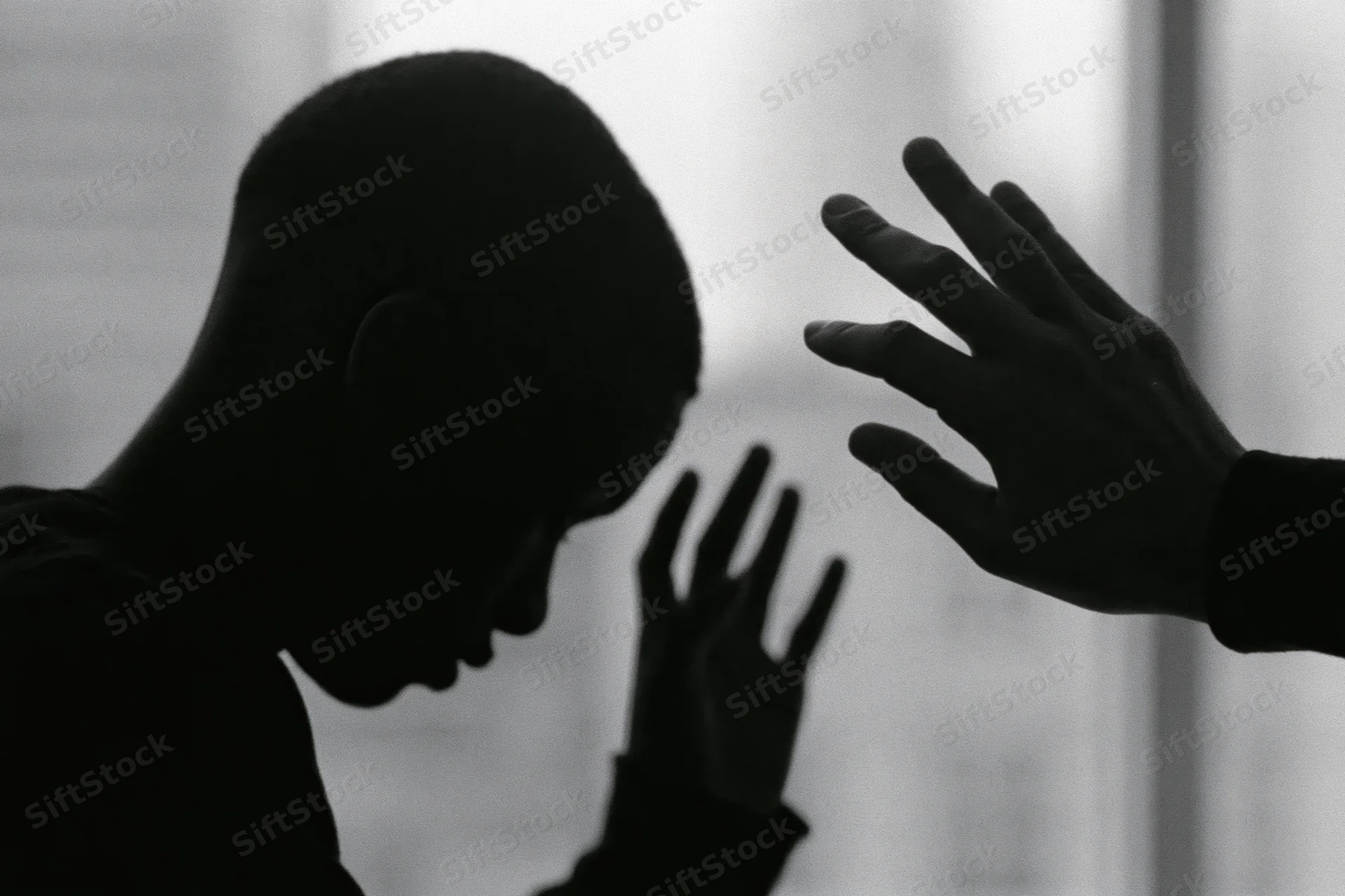 Silhouette of a young black man with head bowed and hands raised in a gesture of distress against a bright window background