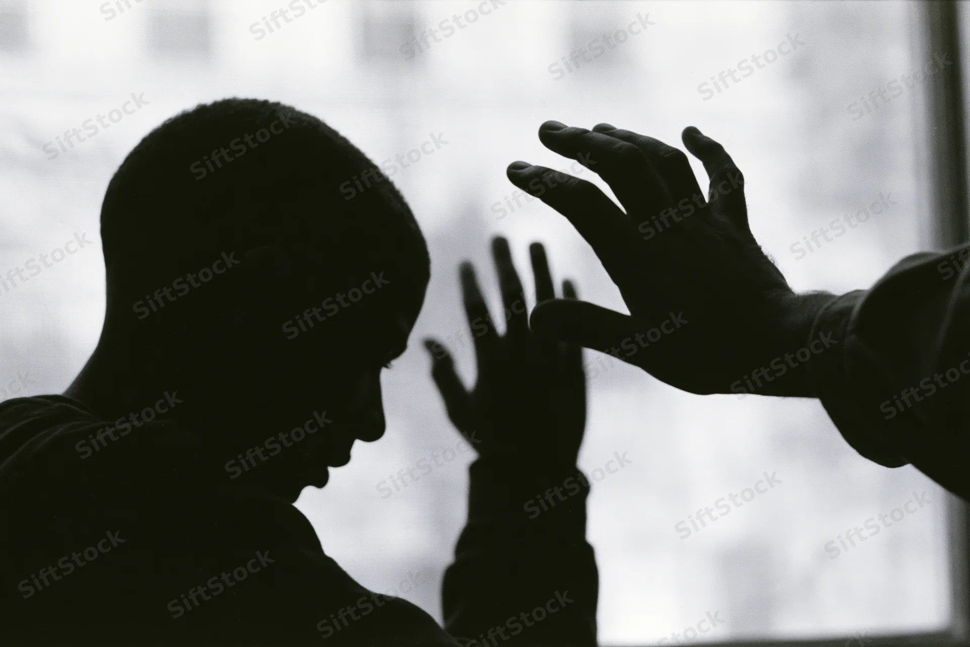 Silhouette of a young man with head bowed and hands reaching toward another hand near a window