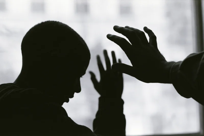 Silhouette of a young man with head bowed and hands reaching toward another hand near a window