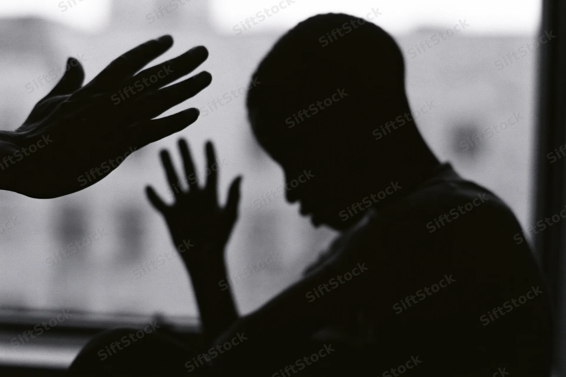 Silhouette of a young person sitting with head bowed and one hand raised toward a reaching hand near a window