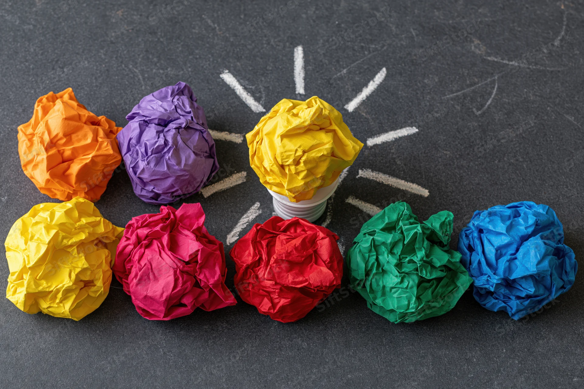 Crumpled colorful paper balls arranged in a circle around a central lightbulb on a dark surface