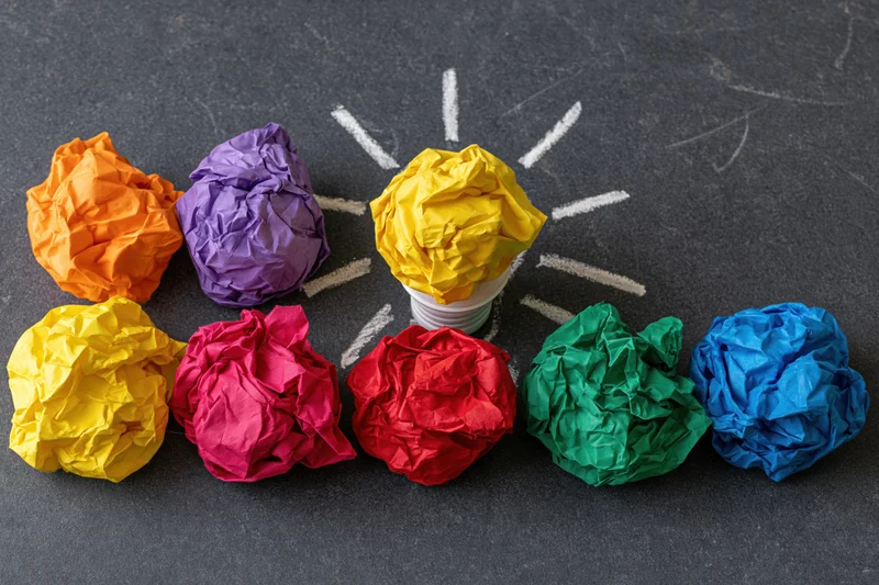 Crumpled colorful paper balls arranged in a circle around a central lightbulb on a dark surface