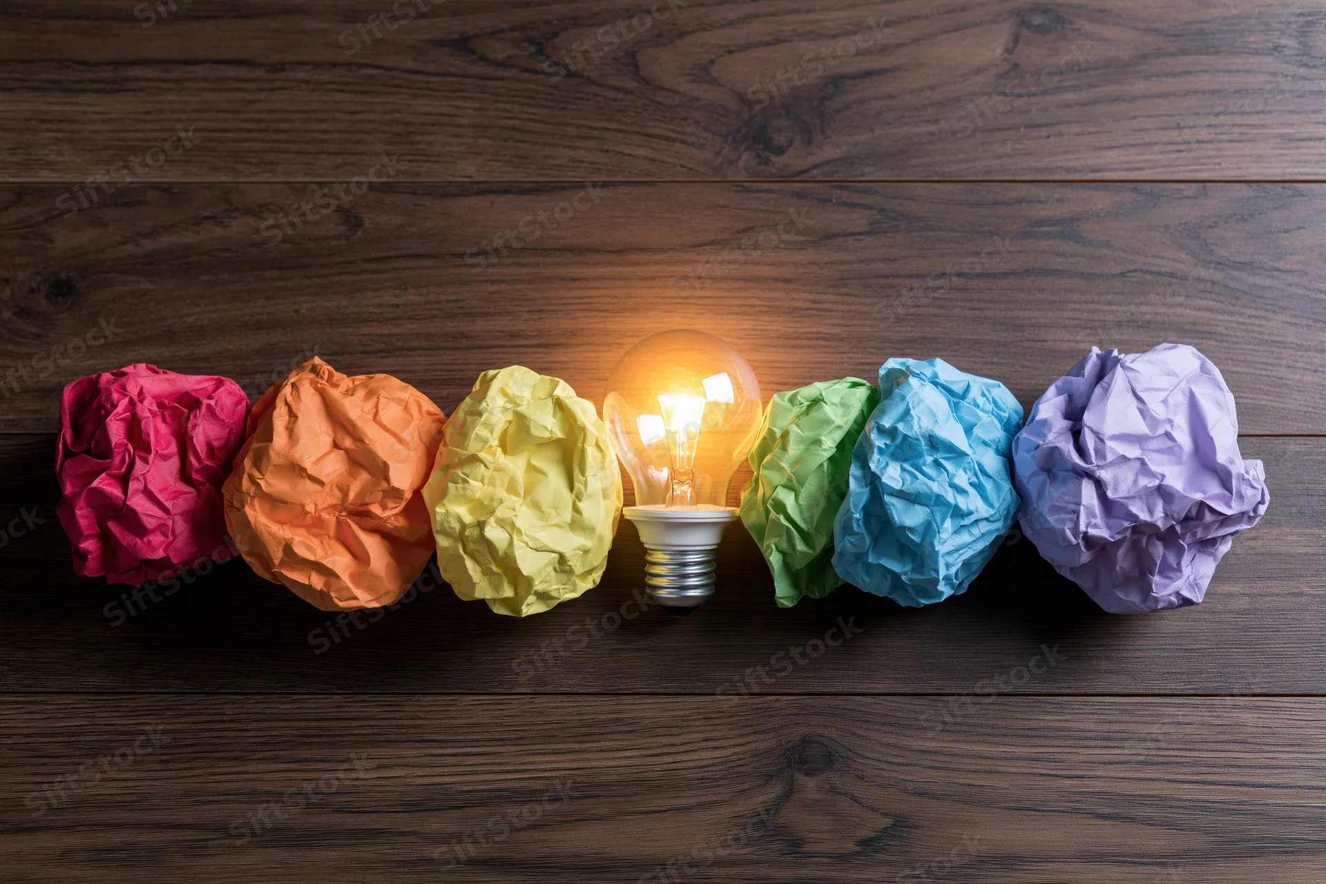 Lightbulb glowing between row of colorful crumpled paper balls on dark wooden surface