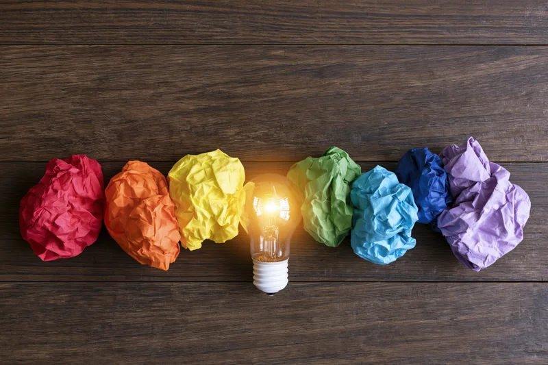 Glowing lightbulb surrounded by rainbow-colored crumpled paper balls on dark wooden surface