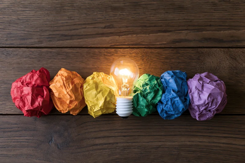 Glowing lightbulb centered among six crumpled rainbow-colored paper balls on dark wooden surface