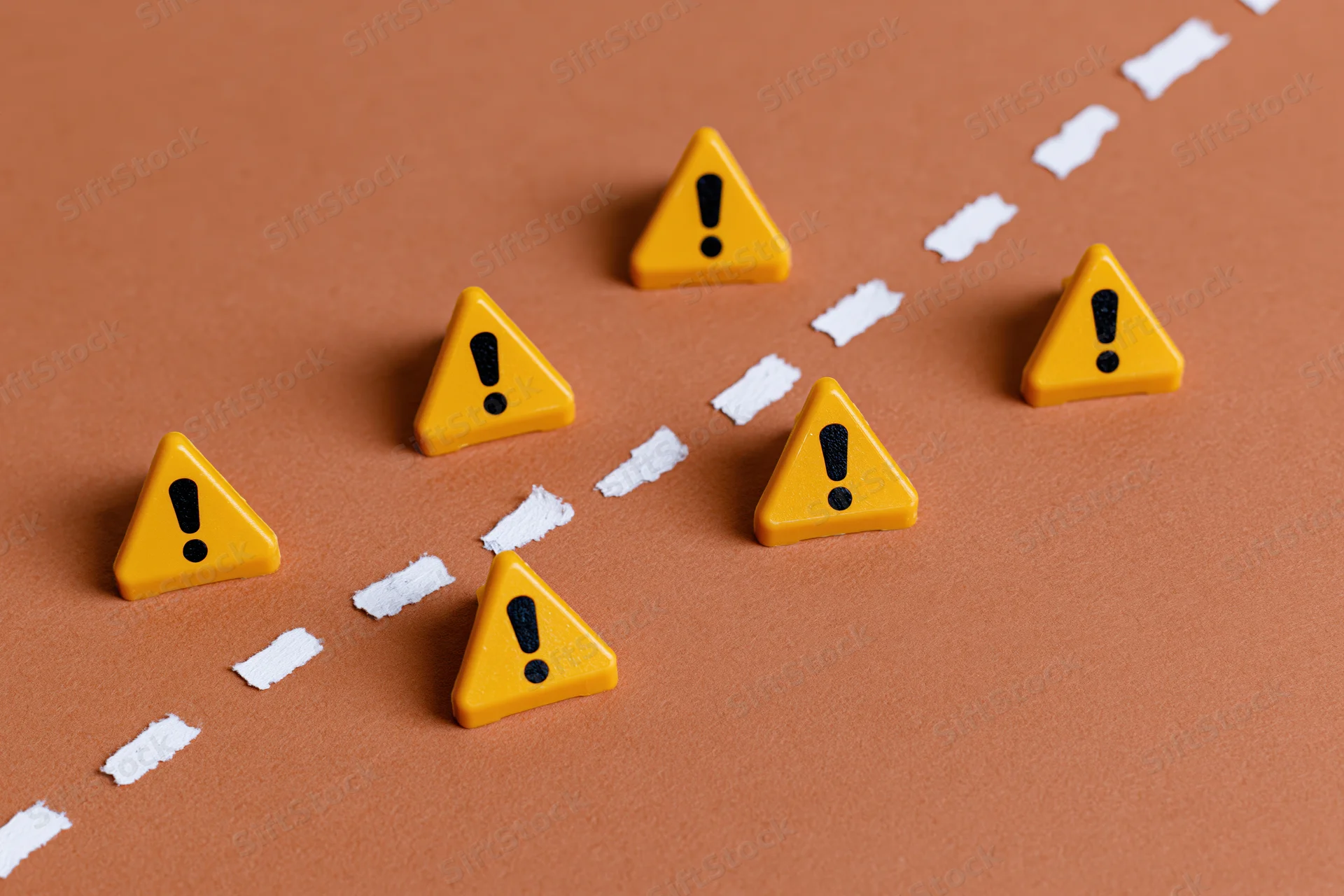 Yellow triangular warning signs with exclamation marks arranged along a dashed white line on a brown surface