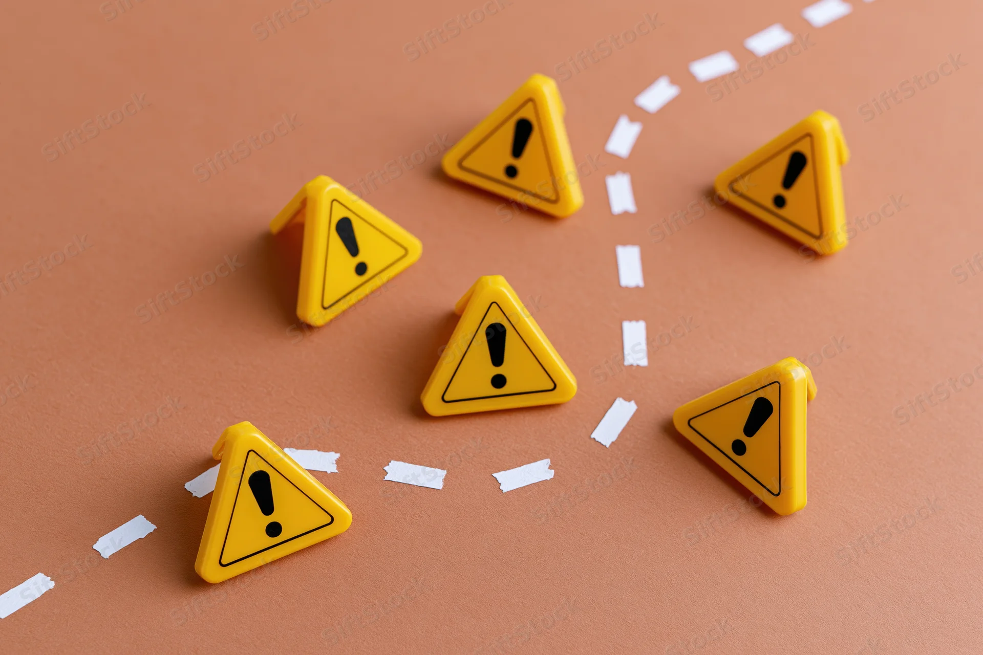Six small yellow triangular warning signs with black exclamation marks arranged around a white dashed path on a brown surface