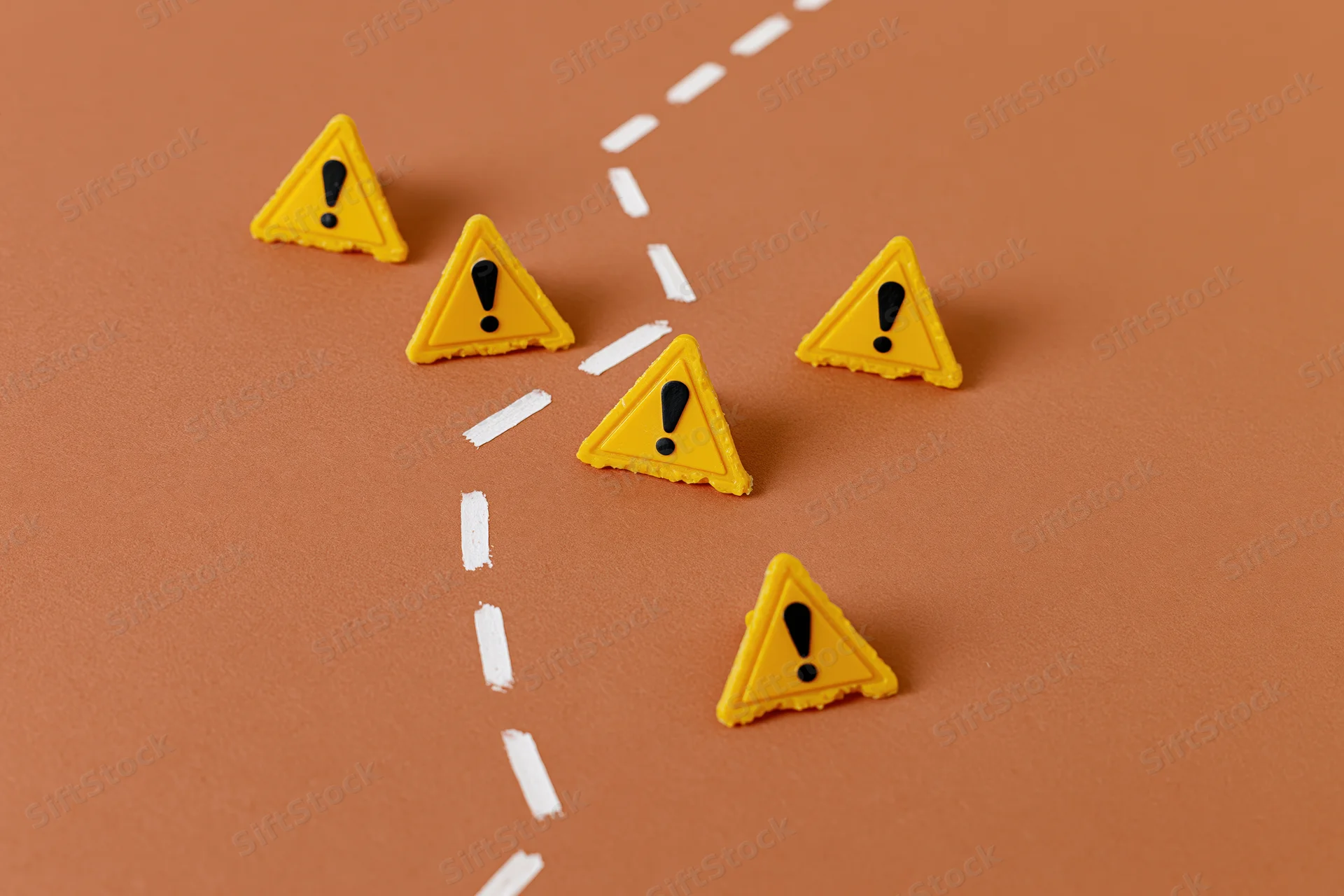 Yellow triangular warning signs with exclamation marks arranged along a dashed white path on a brown surface