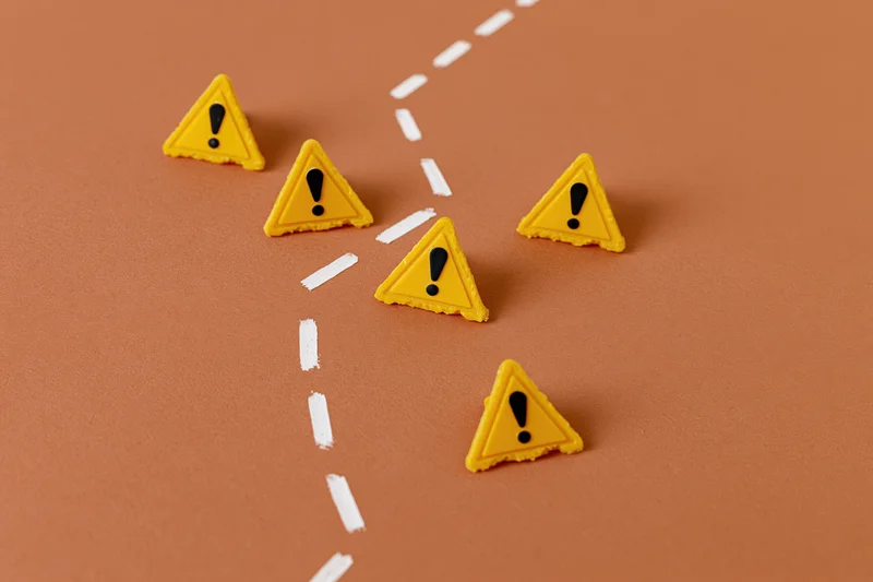 Yellow triangular warning signs with exclamation marks arranged along a dashed white path on a brown surface