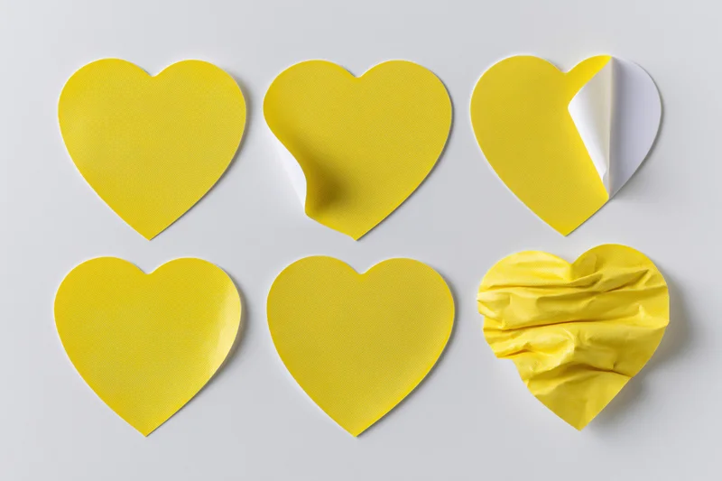 Six yellow heart-shaped stickers arranged in two rows on a white surface, one peeled and one crumpled