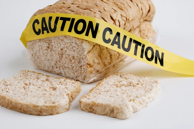 Loaf of sliced white sandwich bread wrapped in plastic with yellow caution tape across it on white background