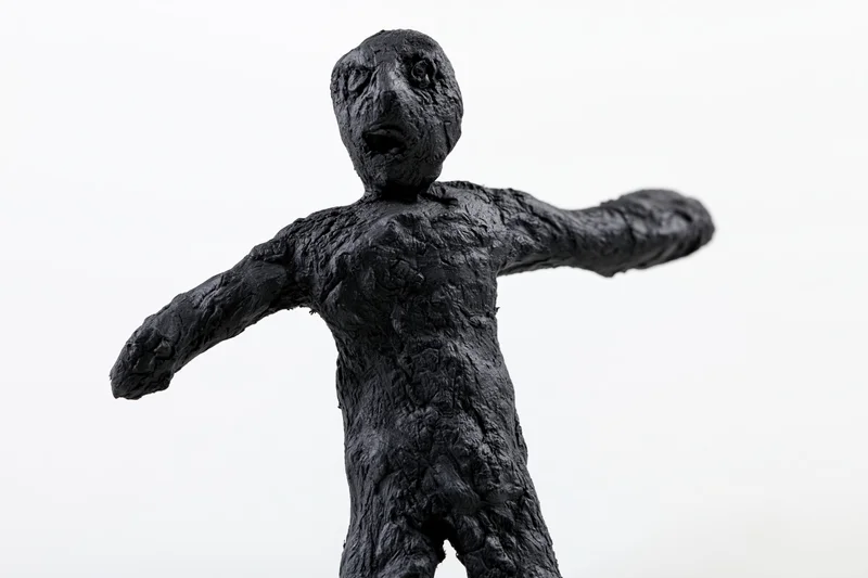 Black clay sculpture of a rough humanoid figurine with arms outstretched, isolated on white background