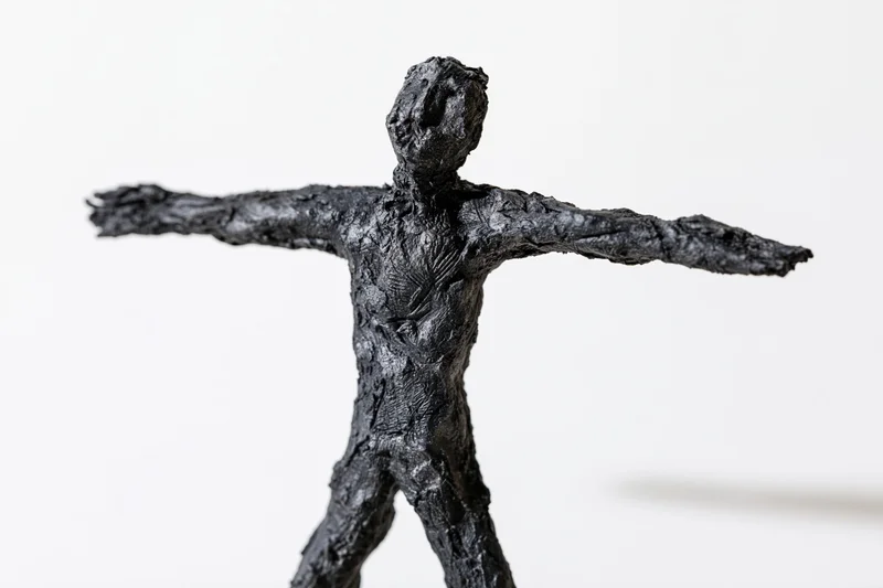 Black handmade clay sculpture of a rough humanoid figure with arms spread standing against white background