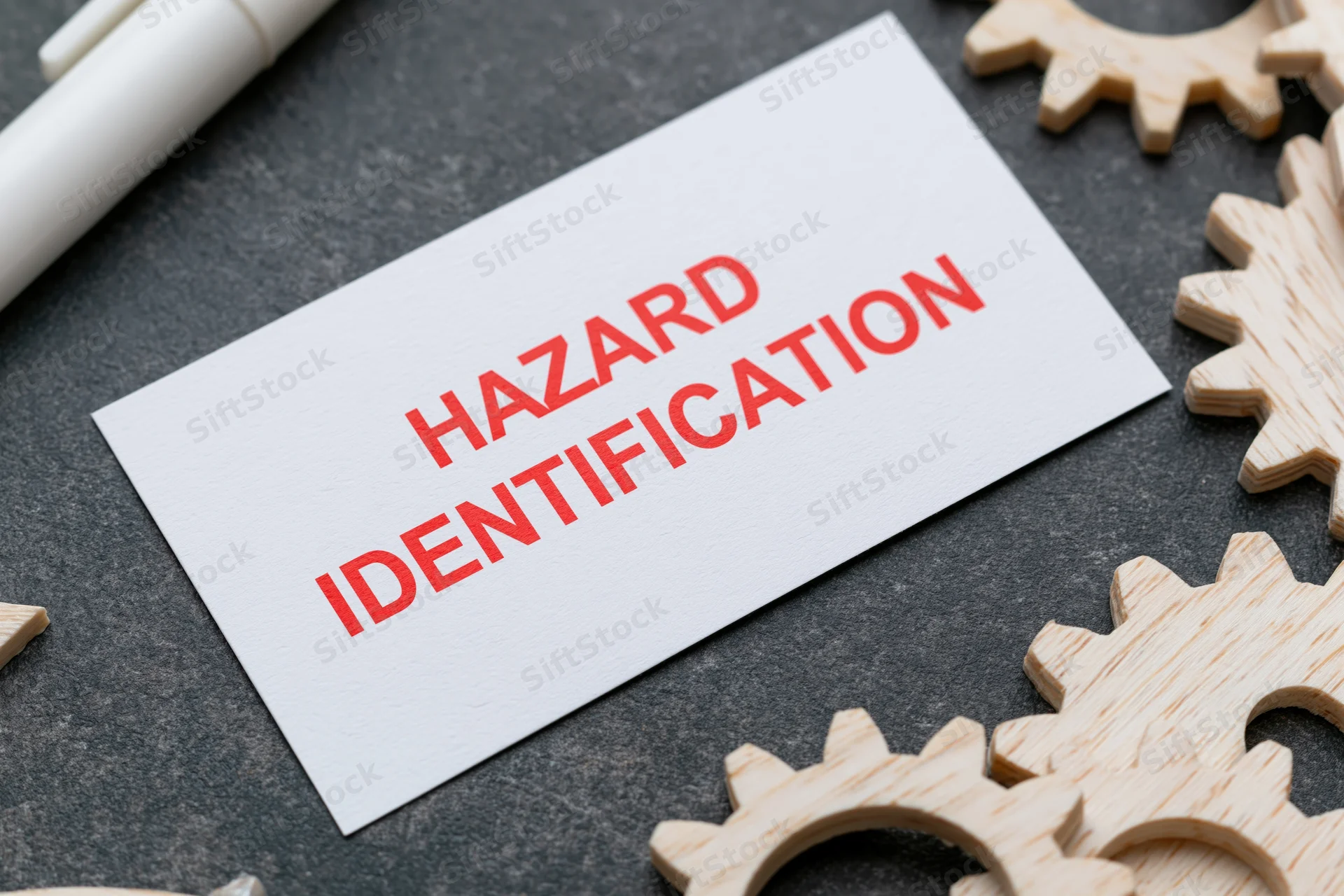 White card with red text hazard identification on dark surface surrounded by wooden gears and a pen