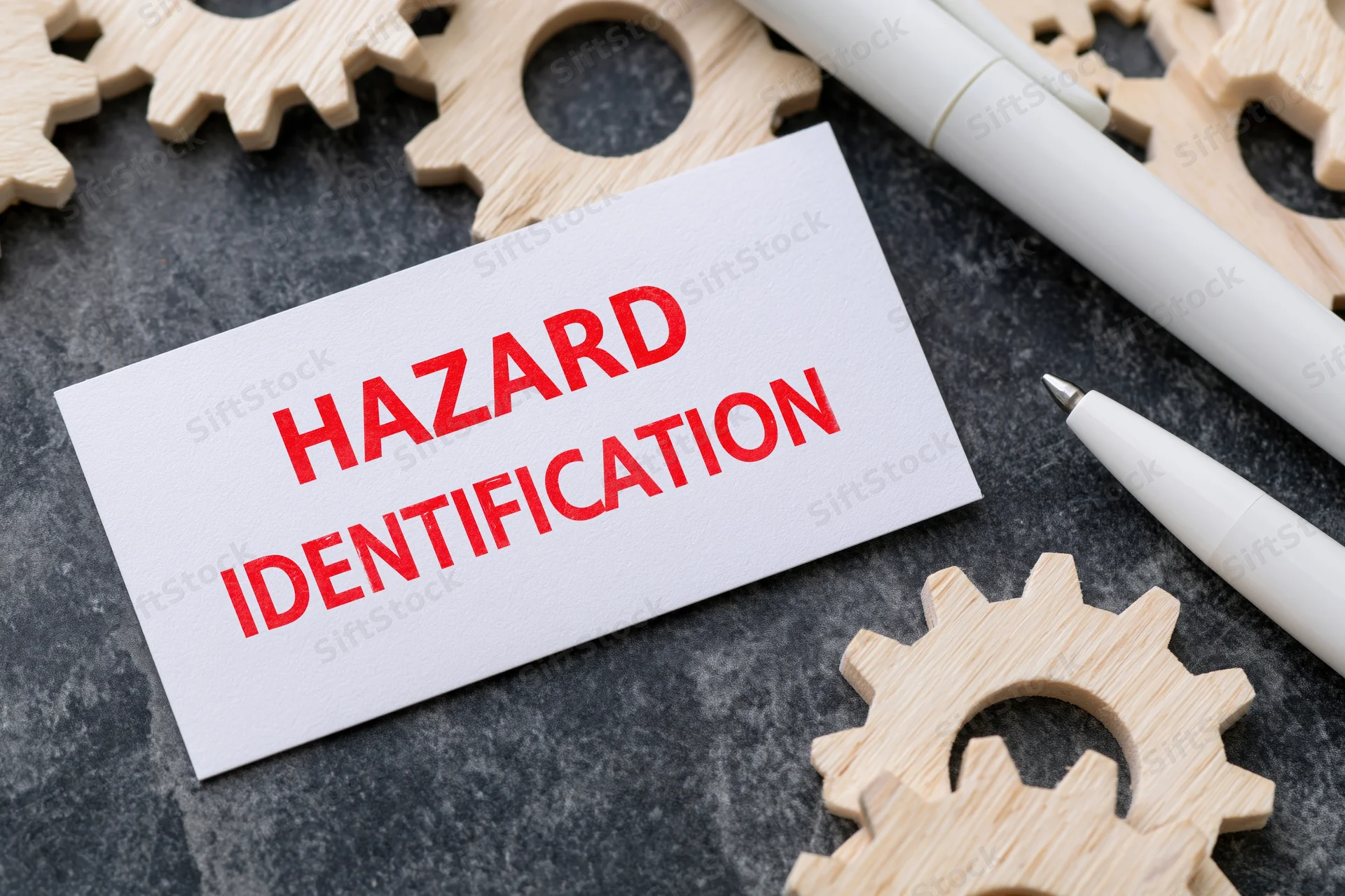White card with red text hazard identification on dark surface surrounded by wooden gears and pens