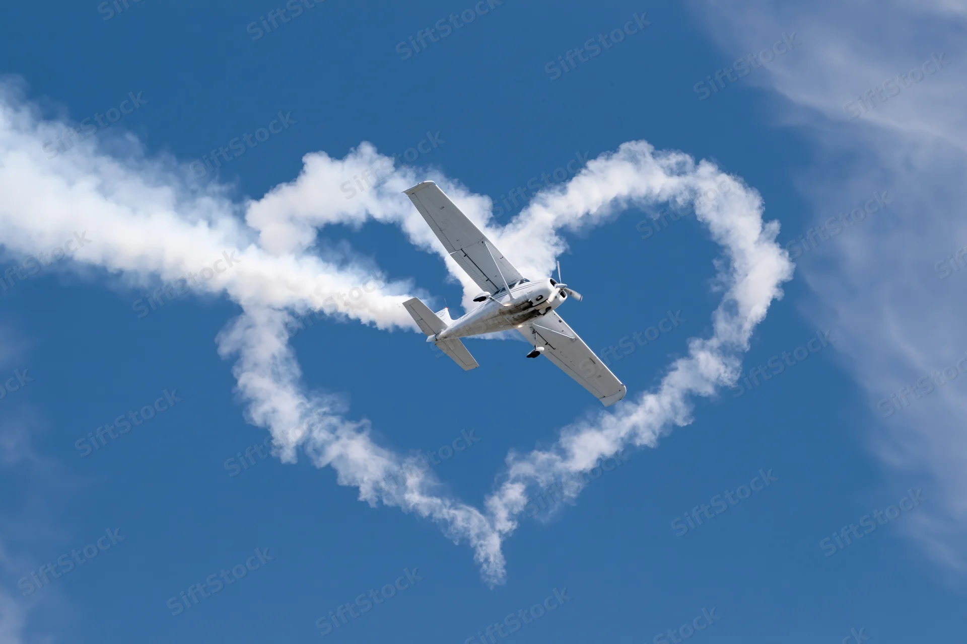 Small white propeller plane flying in blue sky leaving heart-shaped smoke trail