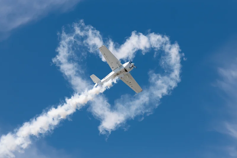 Small white propeller plane flying in blue sky leaving smoke trail shaped like heart