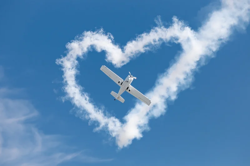 Small white propeller plane flying through clear blue sky leaving heart-shaped smoke trail
